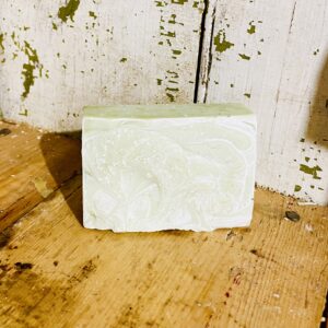 A bar of refreshing acne soap with goat milk, tea tree oil, peppermint oil, and French green clay, displayed against a rustic wooden background.